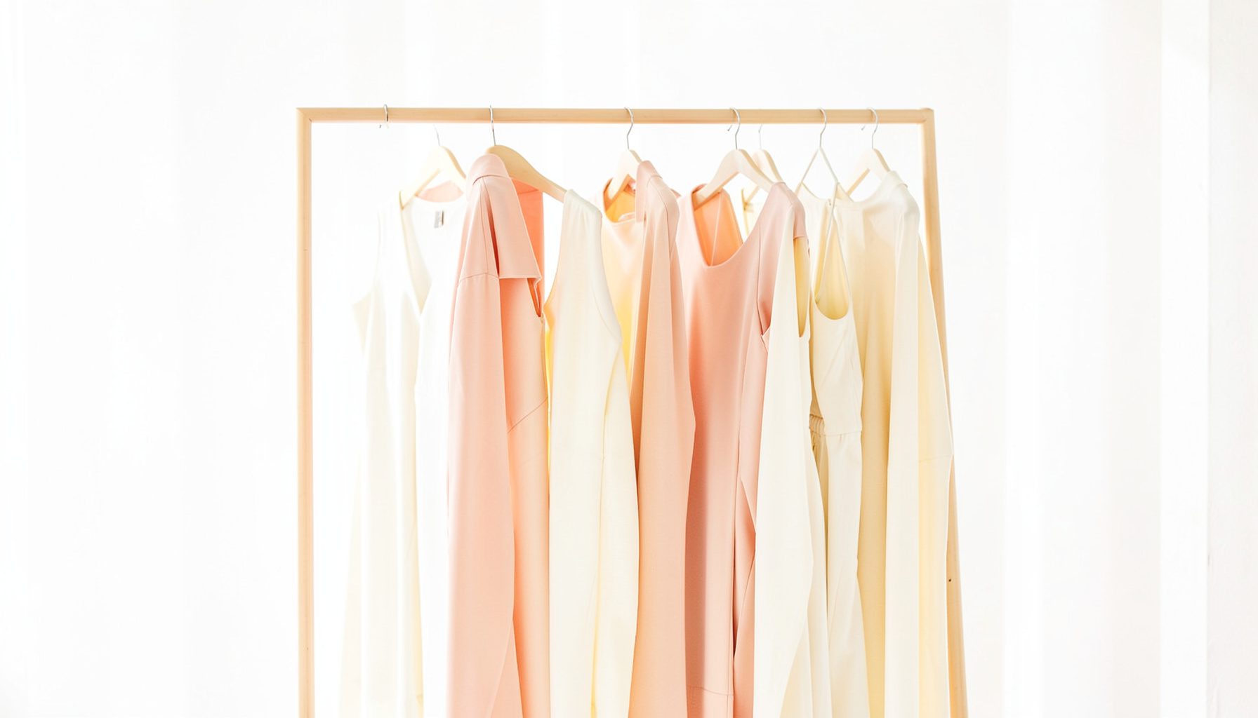 Elegant womenswear pieces in soft cream and blush tones draped on a minimalist rack against a bright airy backdrop