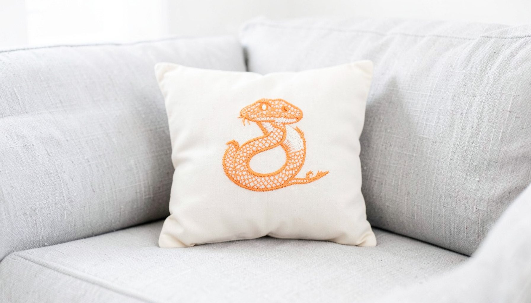 A compact accent pillow with a snake embroidery detail, resting on a linen sofa, soft grey hues