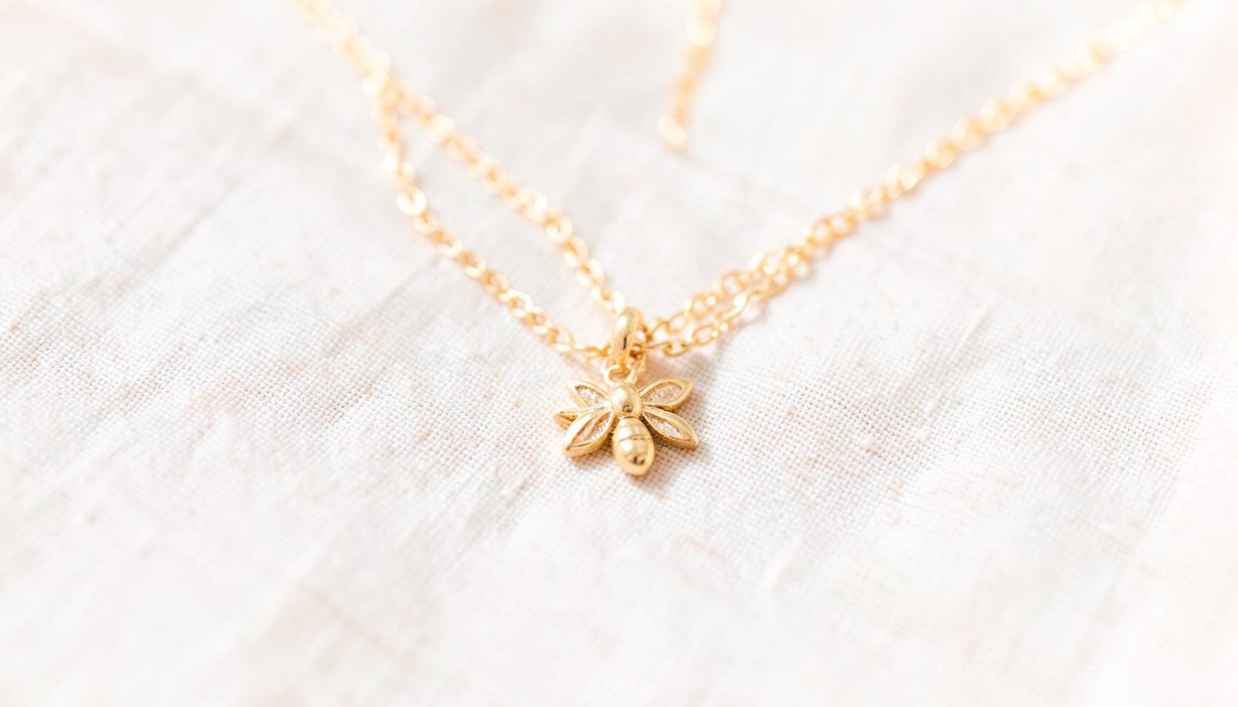 A delicate gold-tone bee charm necklace resting on a pale linen surface, catching soft natural light