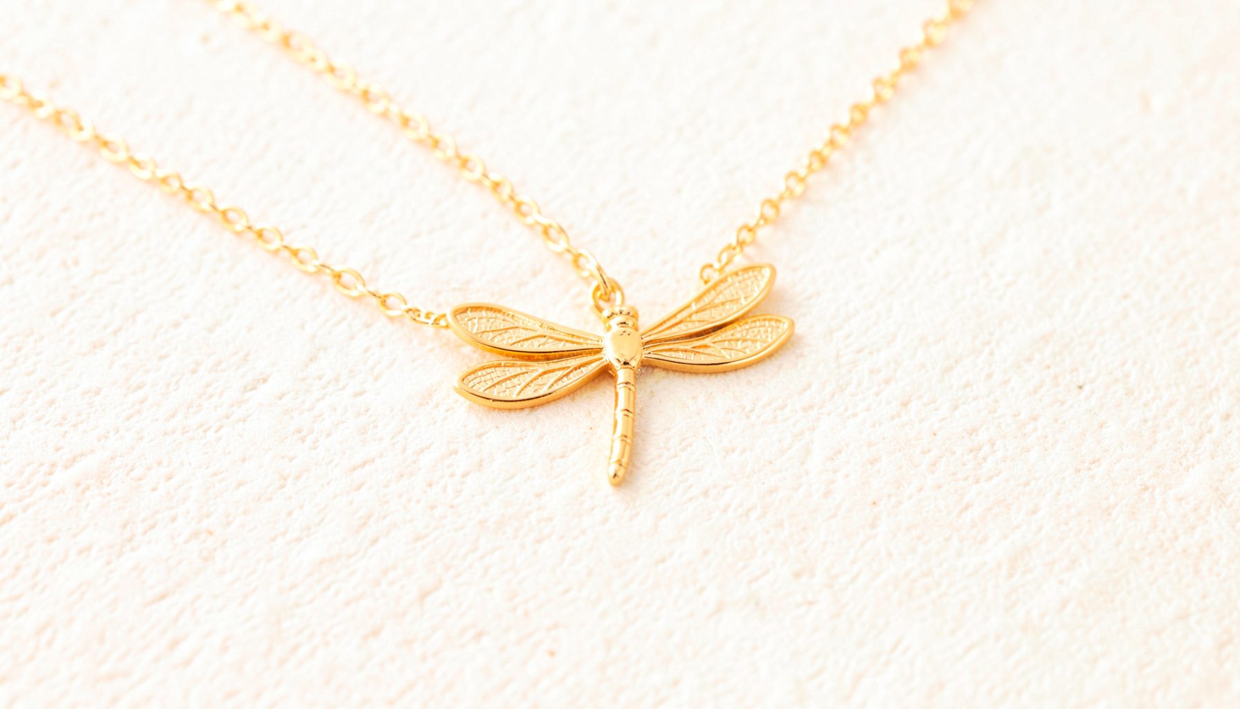 A dainty dragonfly charm necklace in warm gold tones, laid across a textured cream background