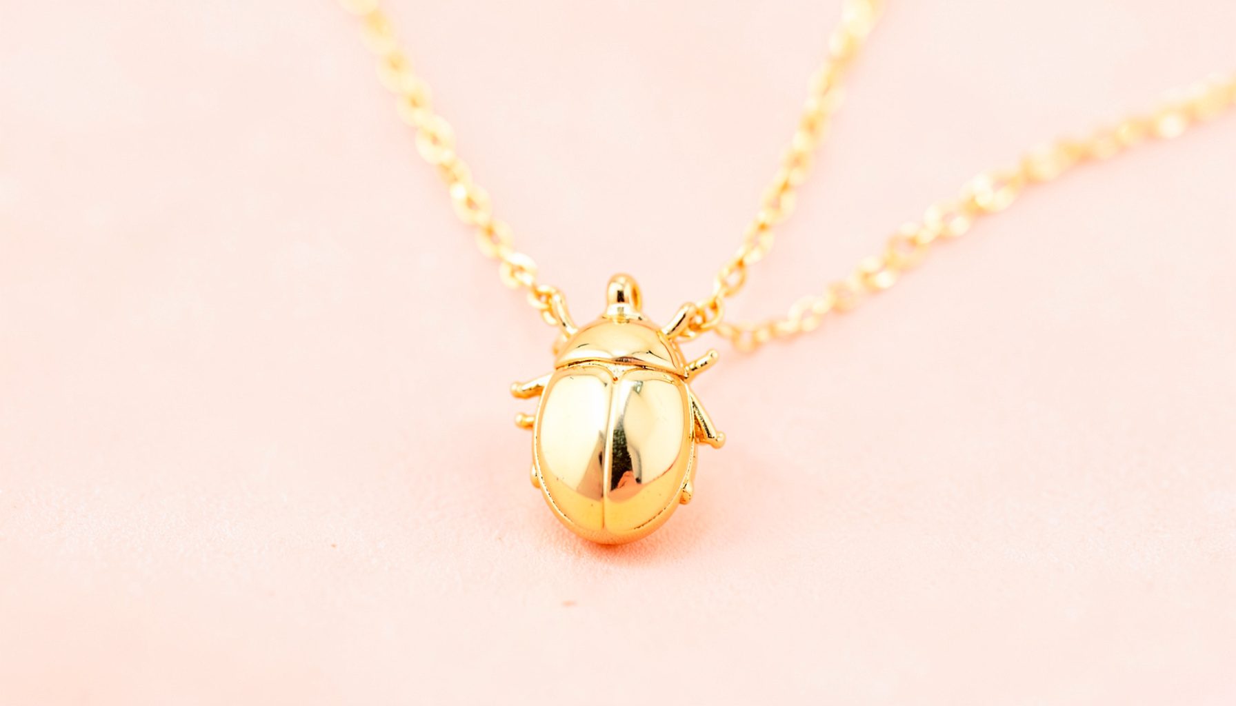 A polished beetle-shaped charm necklace in gold finish, shown against a soft blush-toned backdrop